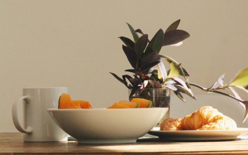 A bowl of fruit on a table with a white mug and plant